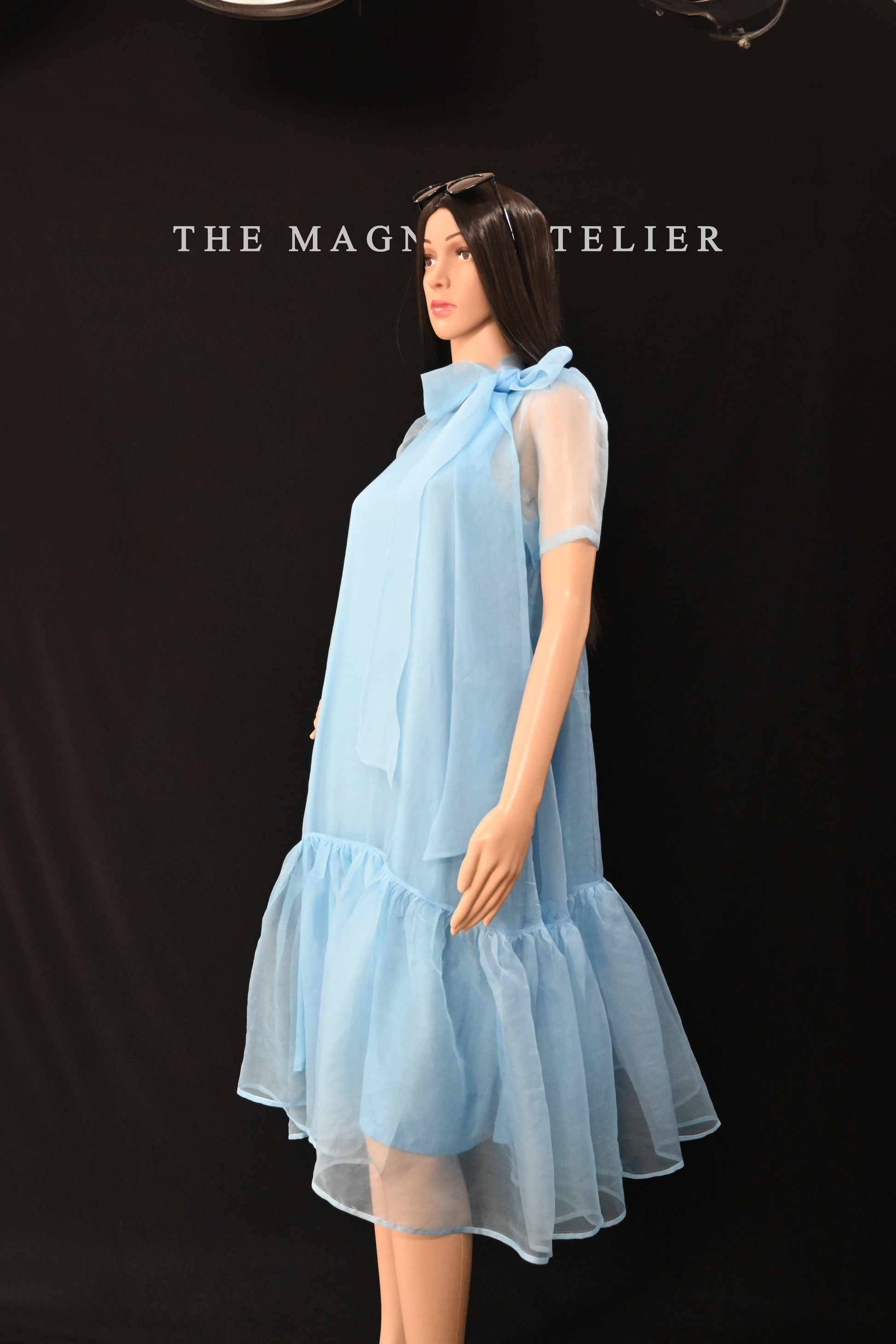 Mannequin in sheer blue organza dress with puff sleeves, ruffled hem, and long neck bow – stylish transparent designer dress for women